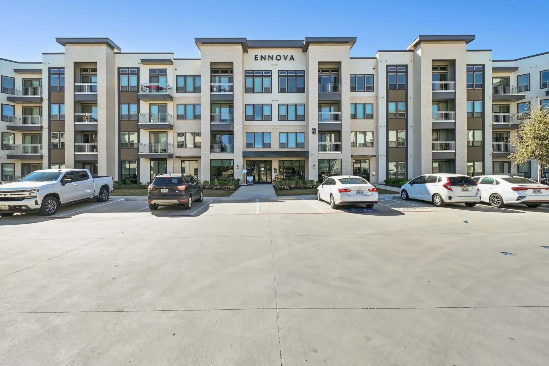 Front of a modern apartment building with balconies and a parking lot in front.