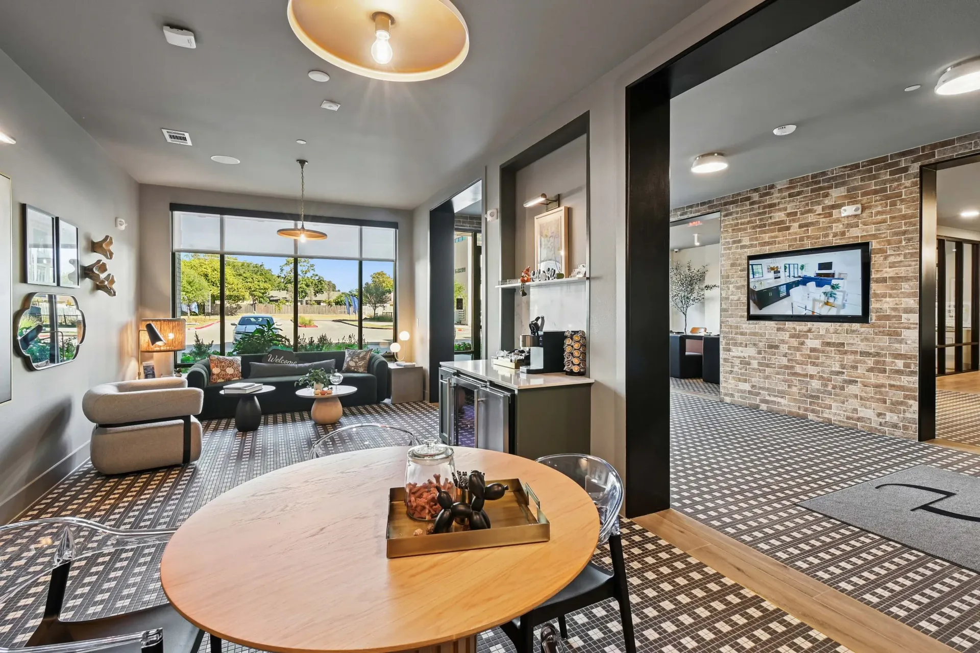 Modern apartment lobby with seating, a round table, large front window, and brick accent wall with a mounted TV.