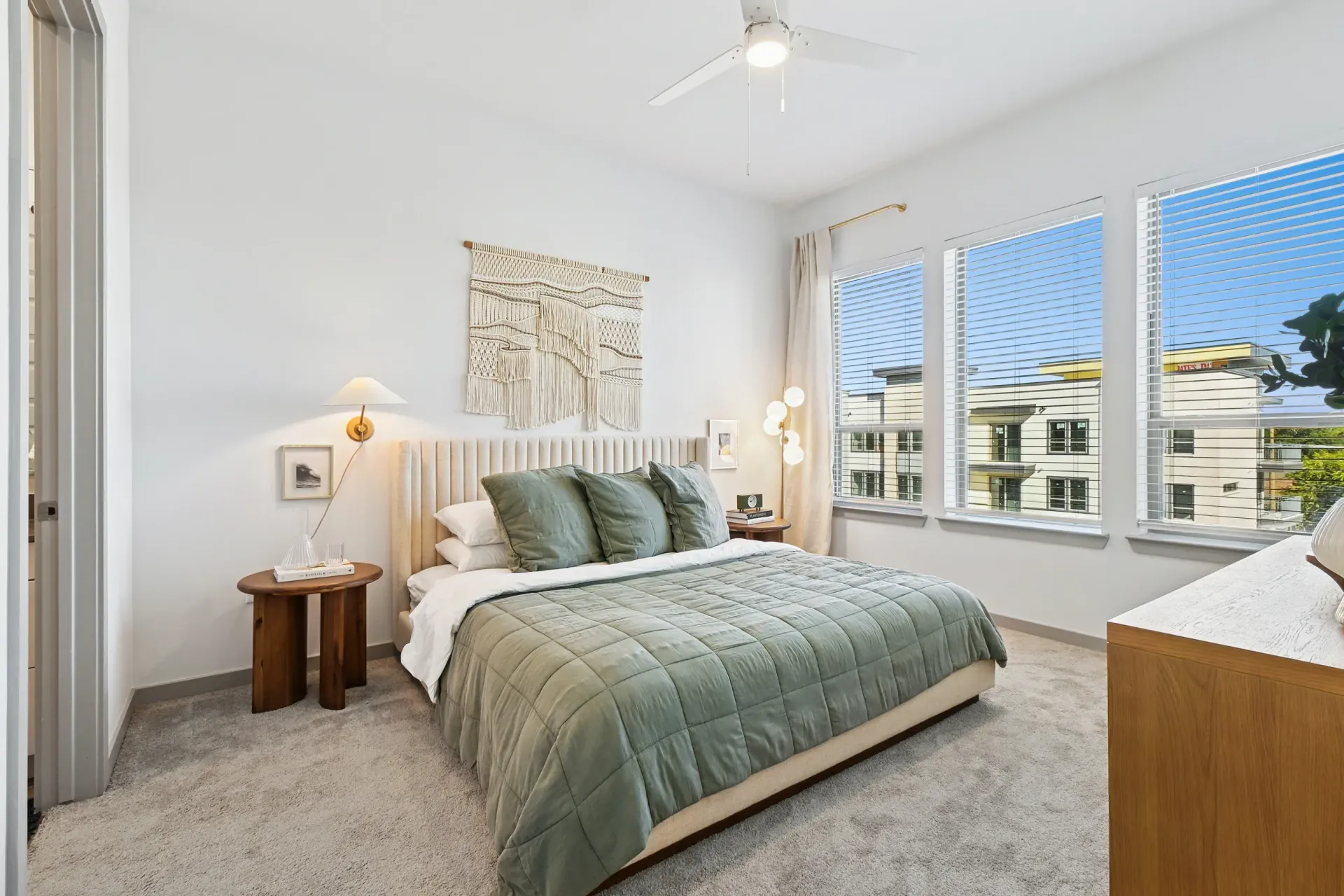 Bedroom with queen bed, upholstered headboard, green quilt, two round wooden nightstands, and large windows