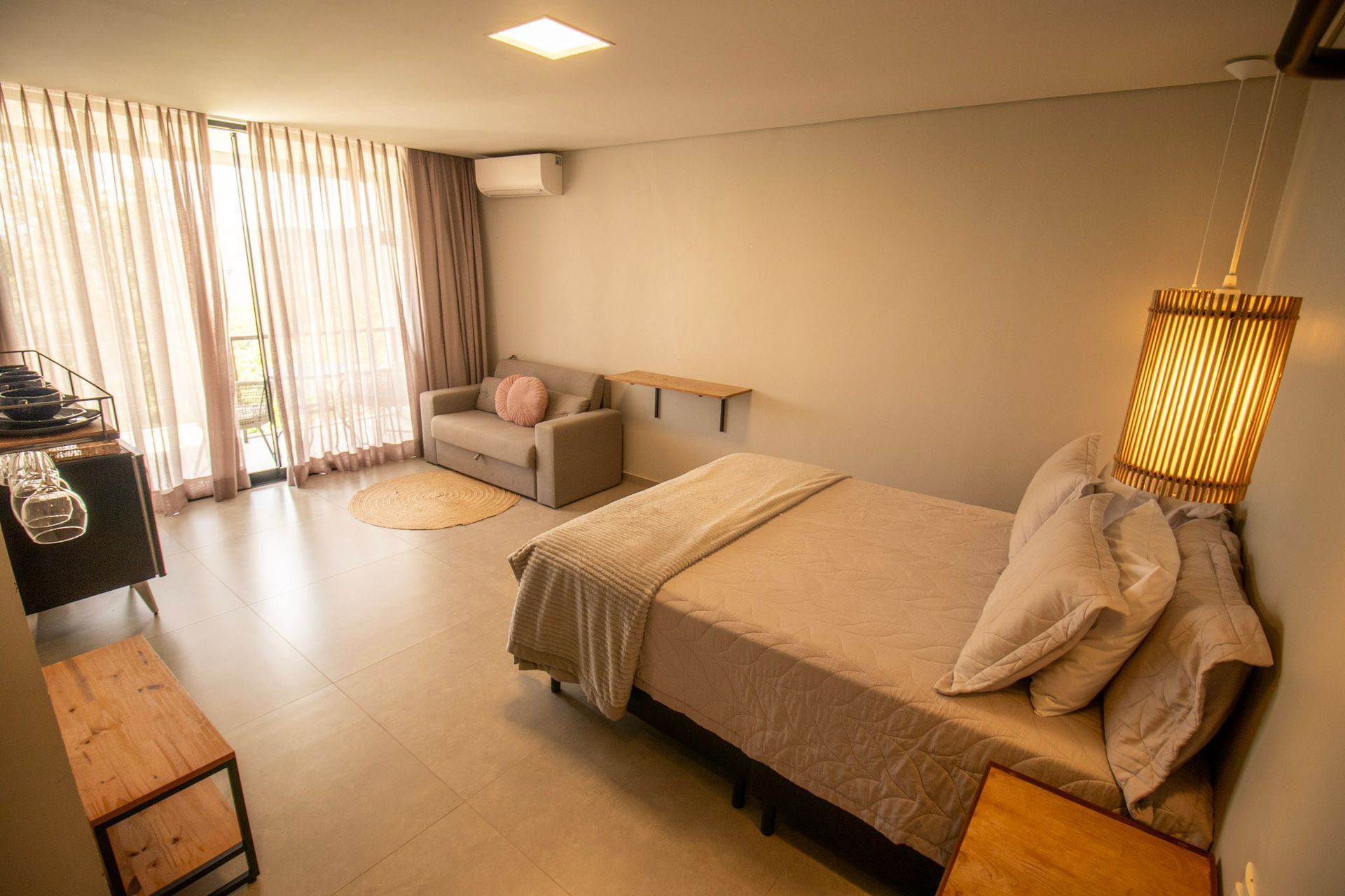 Quarto com cama, sofá pequeno, janela, detalhes em madeira e luminária pendente.