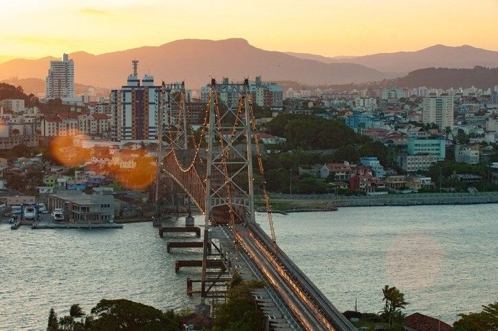 Ponte suspensa sobre a água ao pôr do sol, prédios da cidade ao fundo, luz alaranjada.