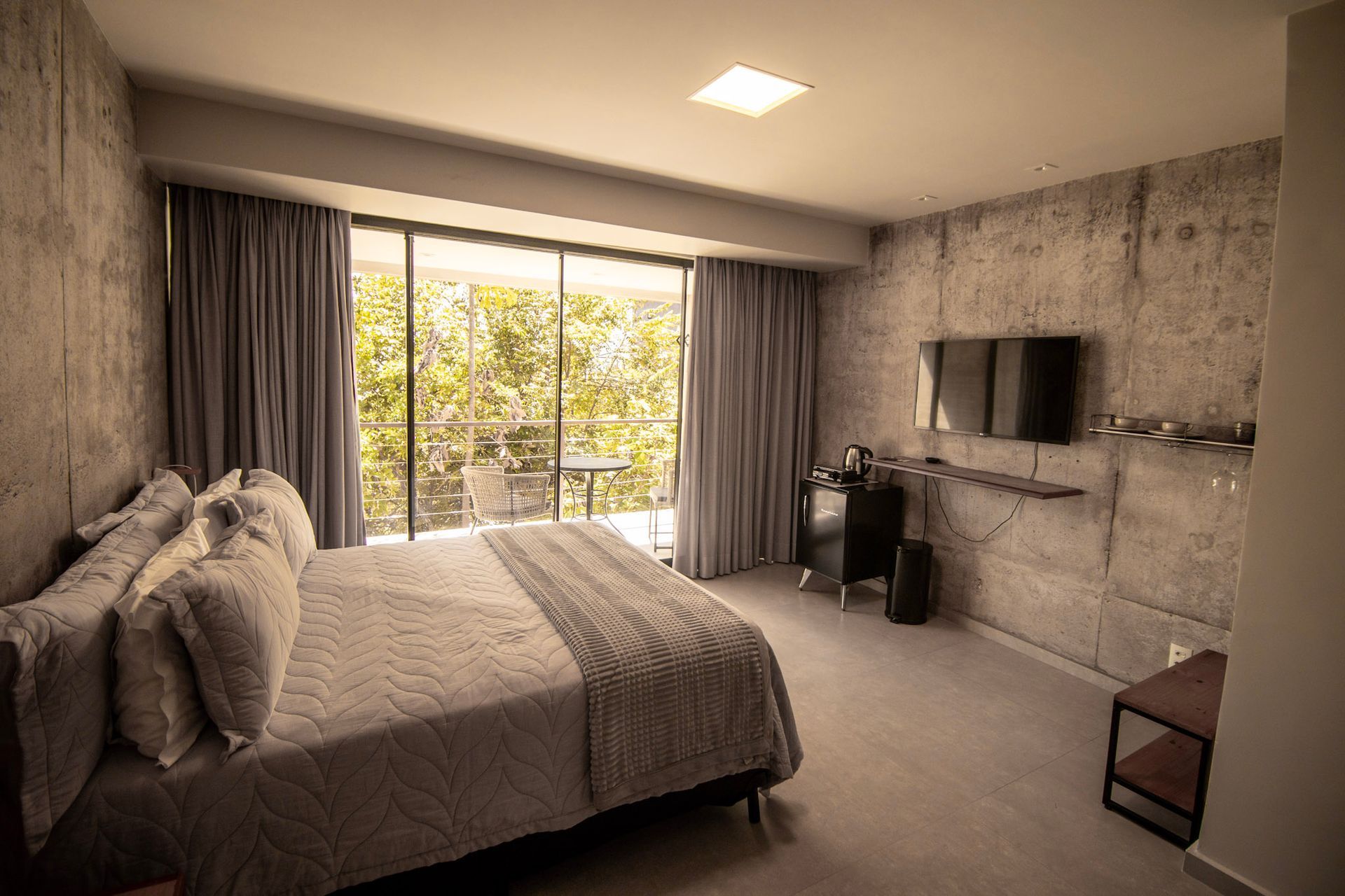 Quarto com paredes de concreto, cama, vista para a varanda e televisão.