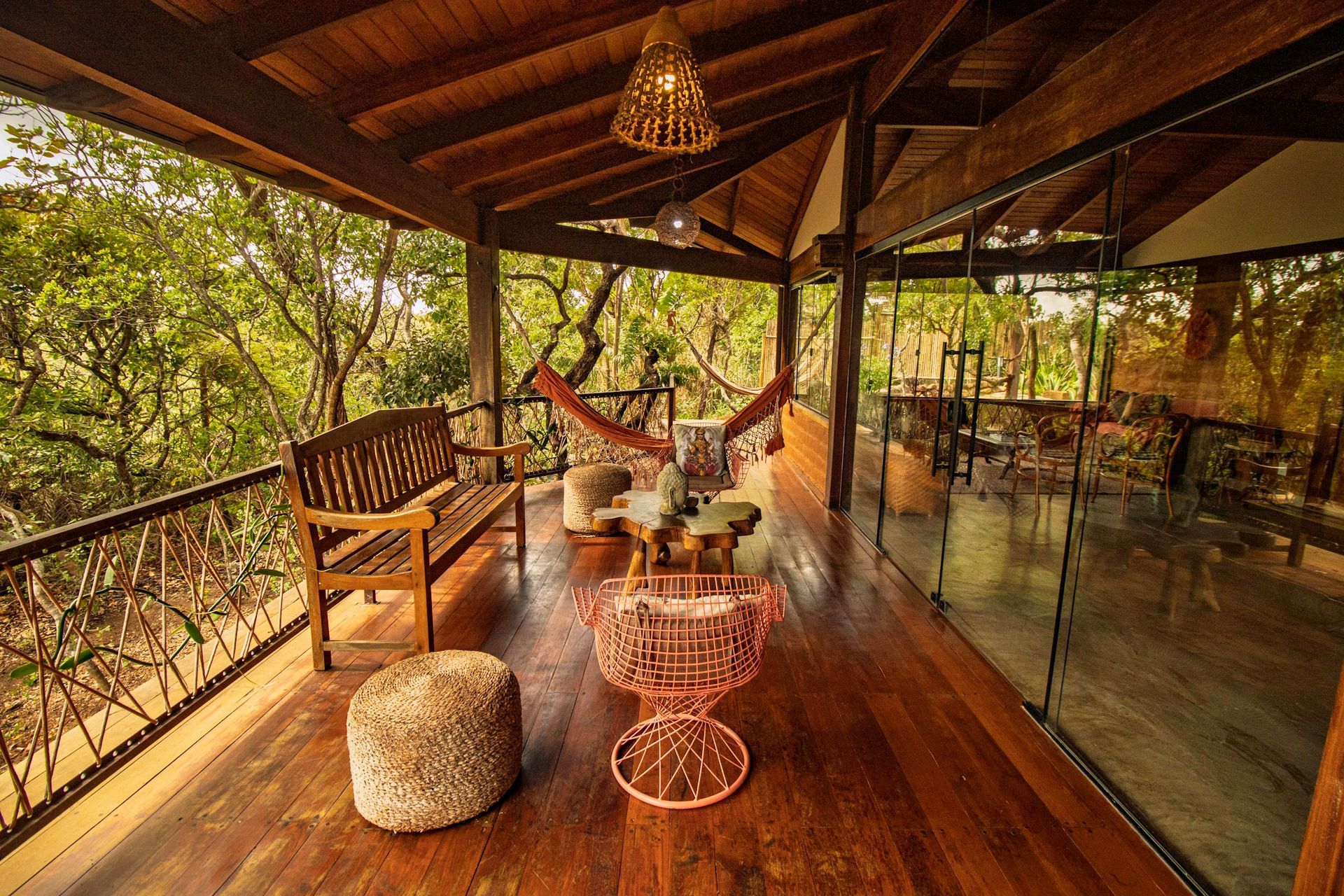 Varanda de madeira com mobília, com vista para uma exuberante floresta verde, paredes de vidro e uma luminária pendente.