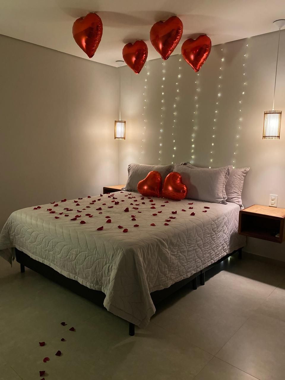 Quarto decorado para o romance com balões em formato de coração e pétalas de rosa sobre a cama. Luzes de cordão e abajures