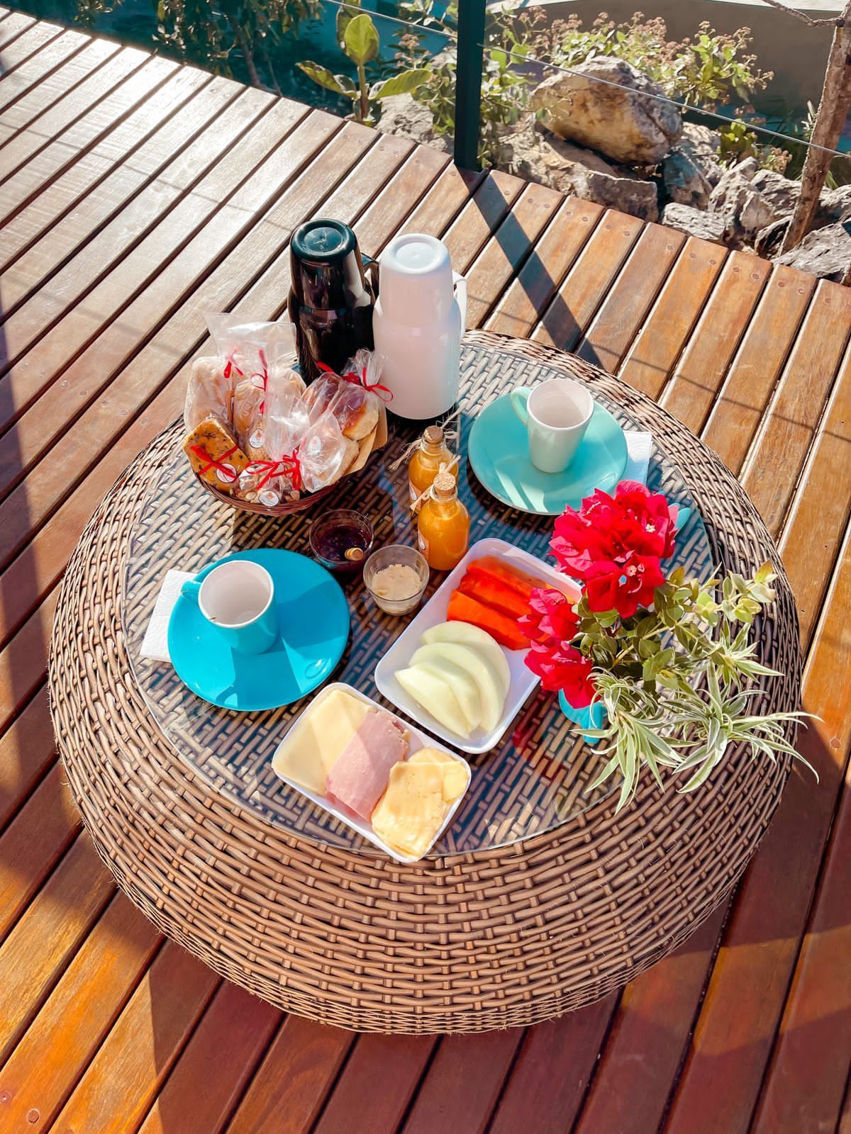 Café da manhã servido em uma mesa de vime ao ar livre. Inclui frutas, pão, bebidas e um arranjo de flores vermelhas.