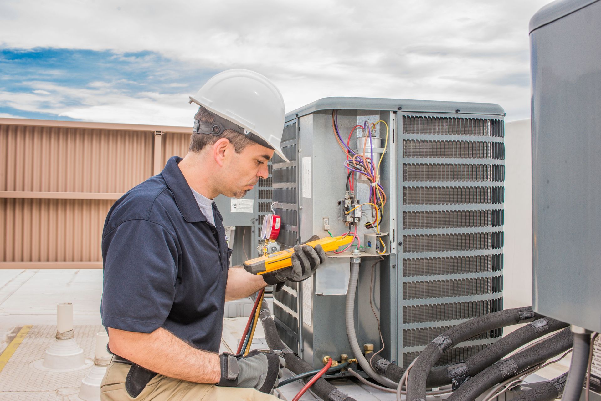 HVAC technician in hardhat uses tools to inspect an outdoor air conditioning unit on a rooftop.