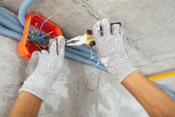 Hands wearing gloves working on electrical wiring in an orange junction box on a concrete ceiling.