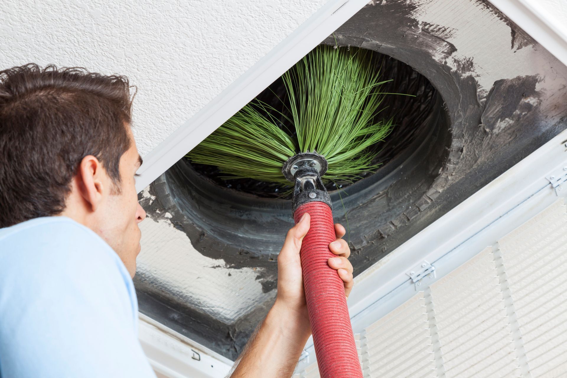 Person cleaning a ceiling air vent with a long, rotating brush.