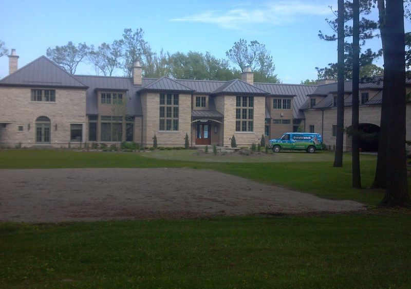 A large, light-colored stone mansion with a dark metal roof, surrounded by a lawn and trees, with a blue van in front.