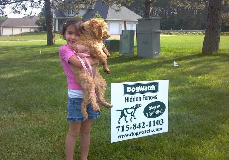 A young child holding a light-brown puppy next to a DogWatch Hidden Fences sign in a grassy yard.