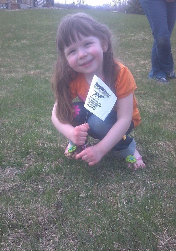 A smiling child crouches on the grass holding a small white flag with a dog logo, wearing an orange shirt and jeans.