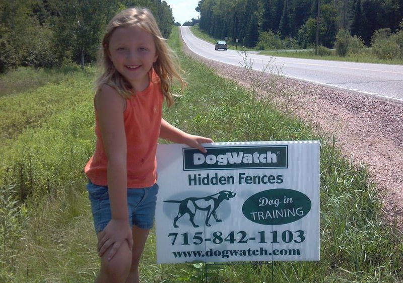 A smiling child holding a DogWatch Hidden Fences yard sign beside a rural road.