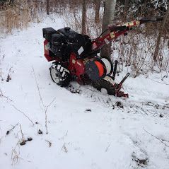 A red motorized trencher sits in the snow-covered woods, positioned next to a tree with an orange cable reel.