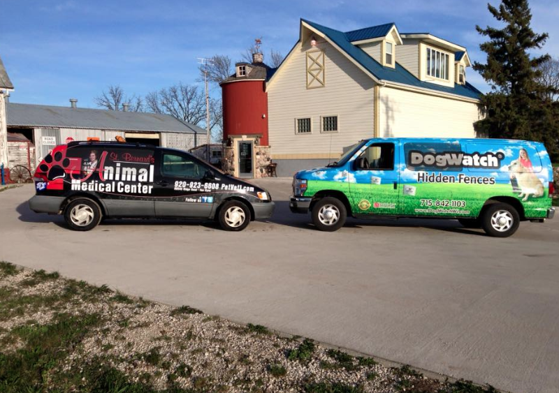 Two service vans parked in a gravel lot in front of a building with a red silo, under a clear blue sky.