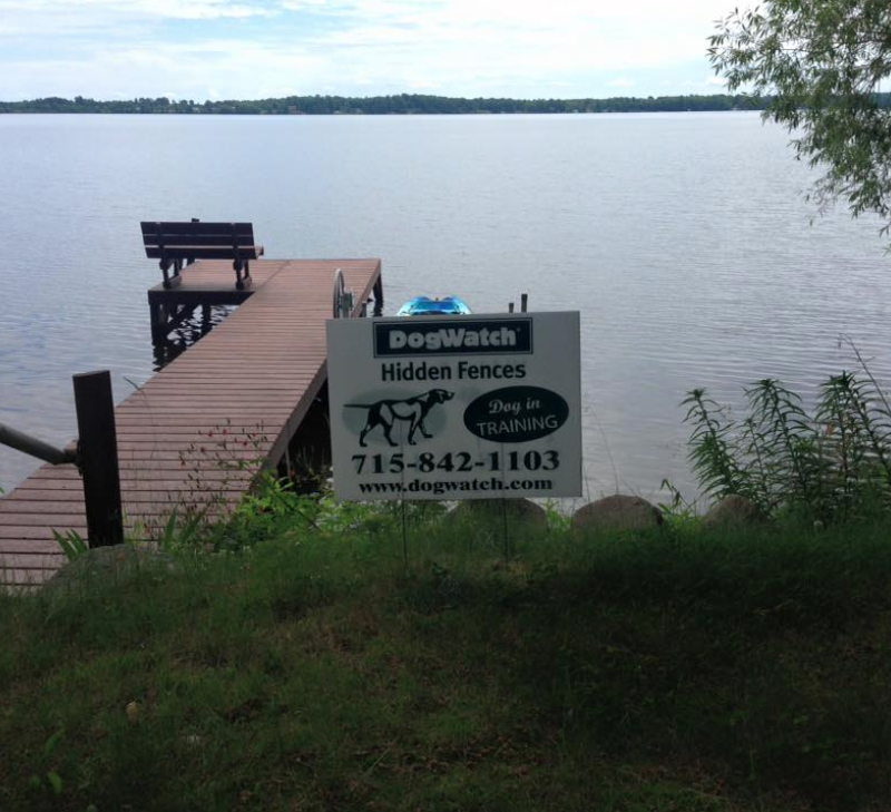 A DogWatch Hidden Fences sign stands on a grassy shore next to a wooden pier extending into a calm lake.