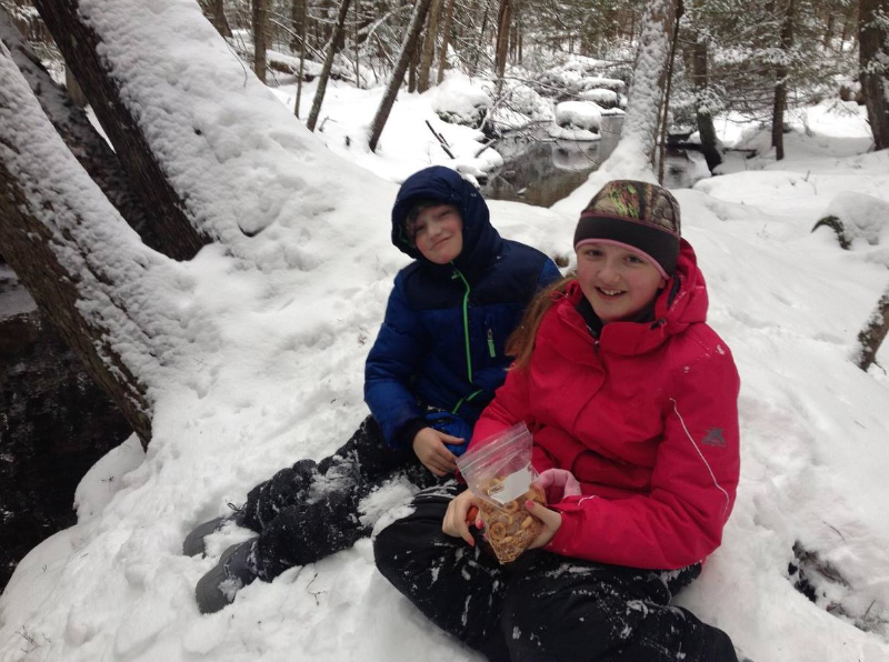 Two children in winter jackets sit in the snow in a forest, holding a plastic bag of snacks.