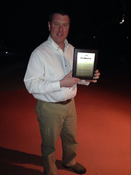 A person stands outdoors at night in a white shirt and khaki pants, smiling while holding a framed award plaque.