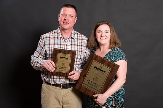 A smiling man and woman stand against a black backdrop, each holding a wooden award plaque.