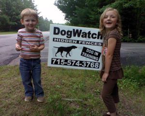 Two children stand outdoors holding a DogWatch hidden fence sign with a dog silhouette and phone number.