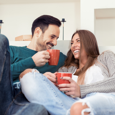 A couple is sitting on an apartment couch, holding a coffee mug, and laughing at each other.