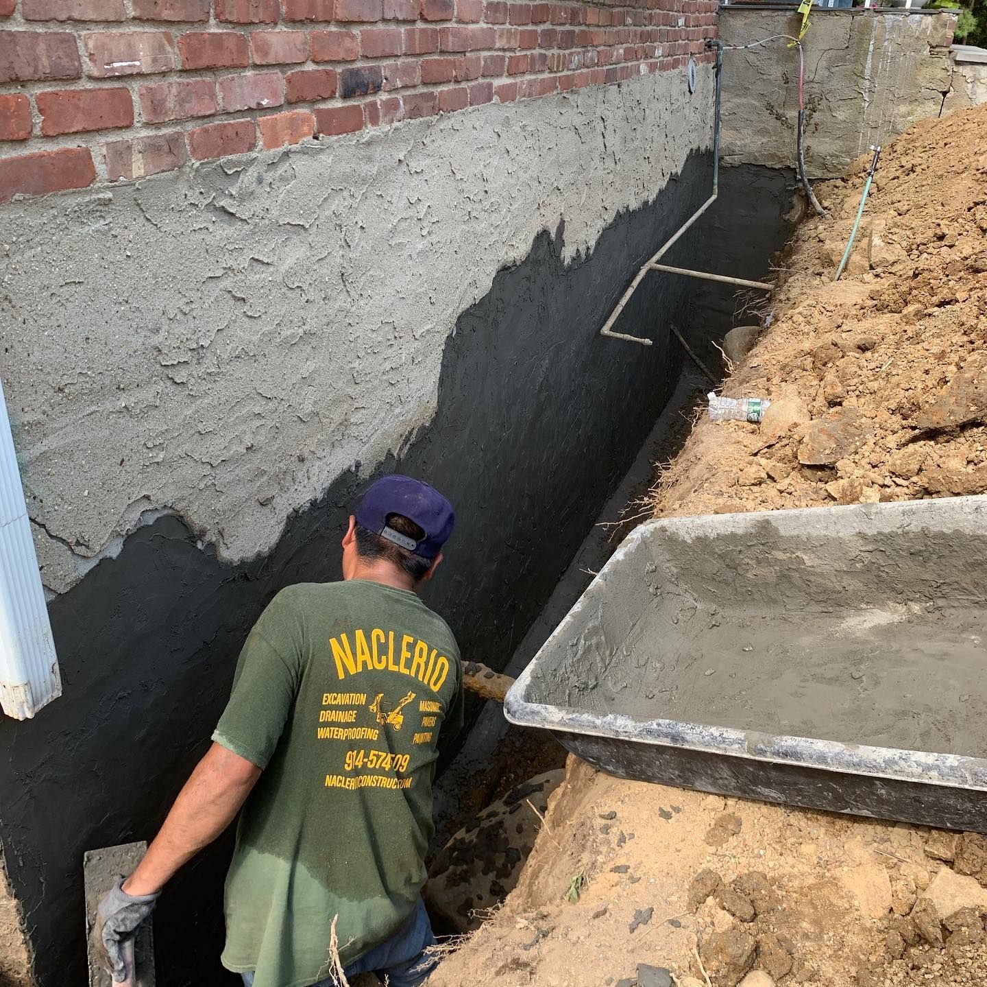 A worker in a green shirt applies black waterproofing material to a concrete foundation wall alongside a trench.