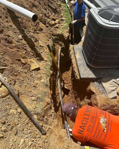 Two workers in a dirt trench next to an outdoor HVAC unit, installing a utility line.