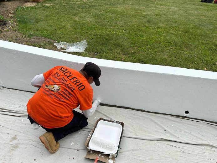 A person wearing an orange shirt kneels on a drop cloth, painting a low white concrete wall with a white paint roller.