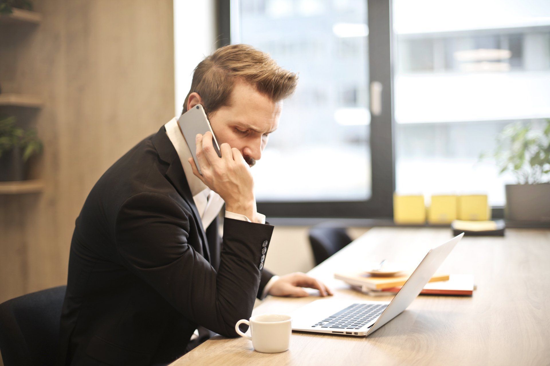 Een man in pak praat op een mobiele telefoon terwijl hij een laptop gebruikt.