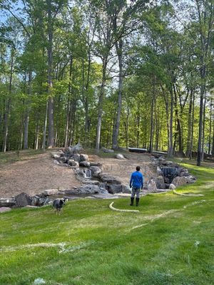 Person in blue jacket standing on grassy lawn near a rocky creek and wooded area.