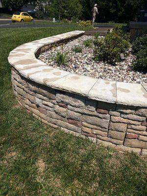 Curved stone retaining wall around a landscaped bed with mulch and shrubs, beside a grassy lawn and street