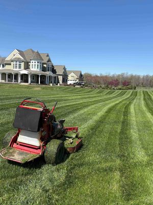 Riding lawn mower cutting striped grass in front of a large house under a clear blue sky