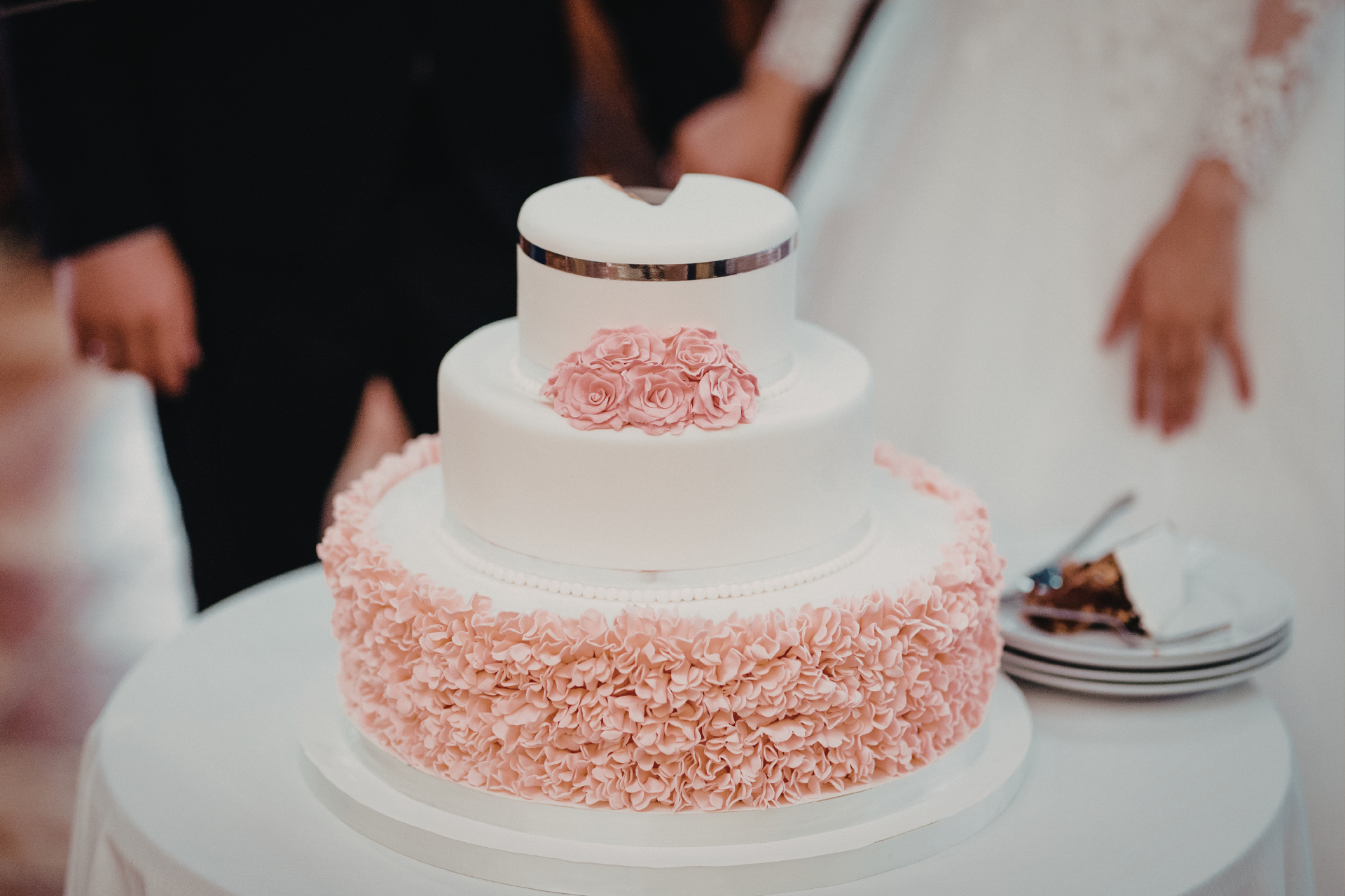 A three-tiered white wedding cake with pink ruffled frosting on the base, decorated with small roses and a silver band.