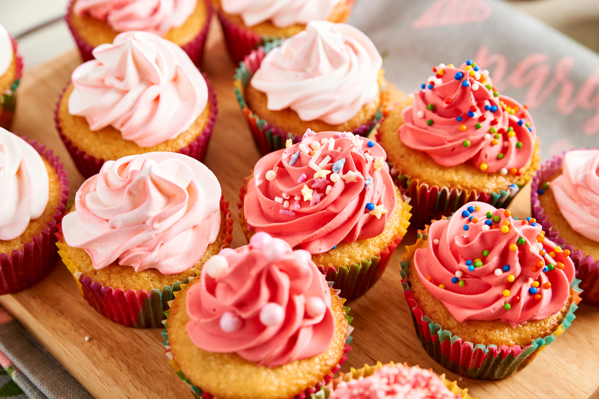 A variety of cupcakes with pink frosting, some topped with colorful sprinkles, arranged on a wooden board.