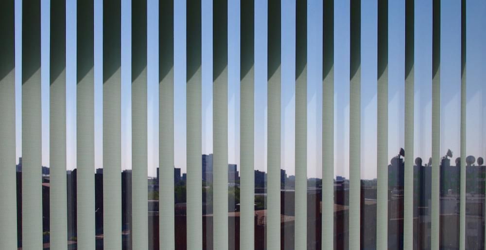 Vertical Blinds Partially Obscuring a Cityscape With Tall Buildings, Casting Shadows. Blue Sky Visible — My Timber Flooring Blinds & Shutters in Erina, NSW