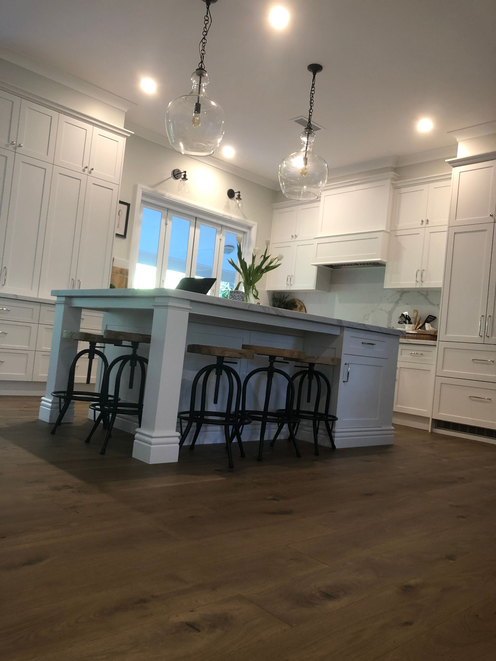 White kitchen with island, pendant lights, and dark wood floors. Black stools at the island. — My Timber Flooring Blinds & Shutters in Erina, NSW
