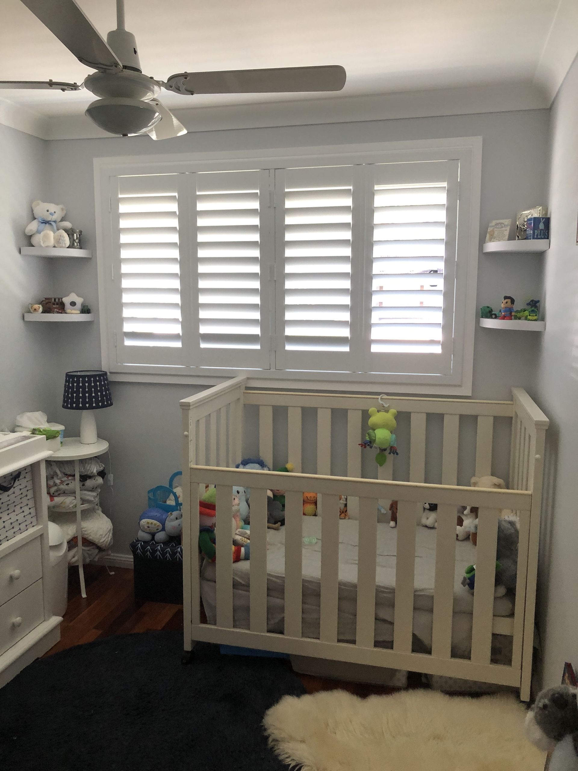 A baby's nursery with crib, dresser, and window with white shutters. Shelves hold toys. — My Timber Flooring Blinds & Shutters in Erina, NSW