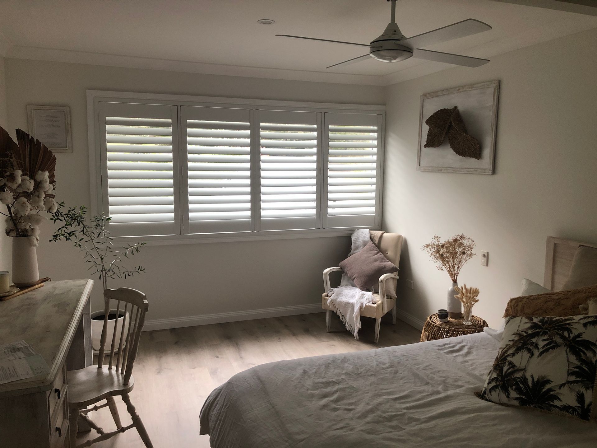 Bedroom with white shutters, chair, desk, and bed. Neutral colours and a horse head artwork. — My Timber Flooring Blinds & Shutters in Erina, NSW