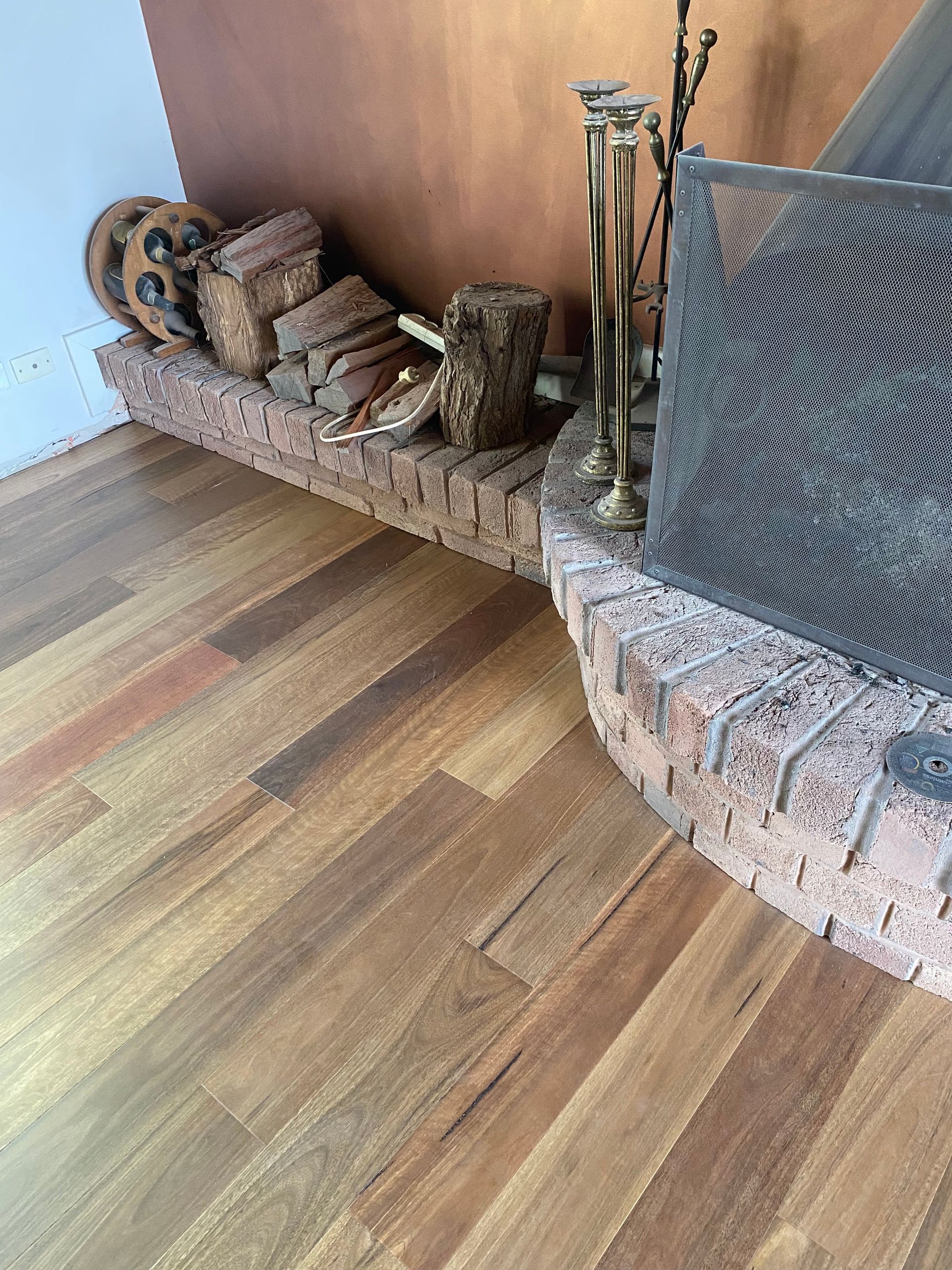 Wood Flooring Next to a Brick Fireplace With a Metal Screen, Firewood, and Bronze Accents — My Timber Flooring Blinds & Shutters in Erina, NSW
