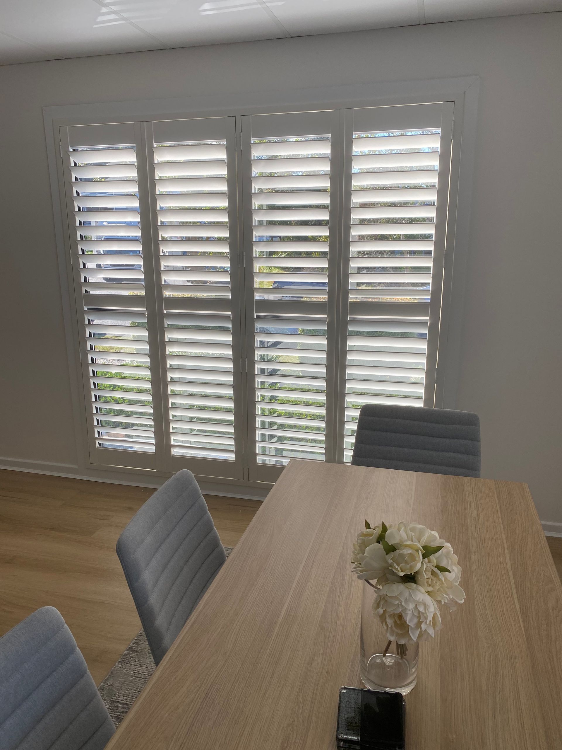 A Dining Room With White Shutters on a Large Window, Wooden Table, Grey Chairs, and a Vase of Flowers — My Timber Flooring Blinds & Shutters in Erina, NSW