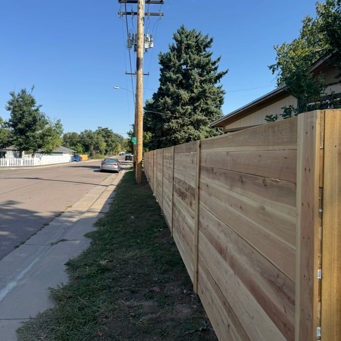 Wooden fence along a sidewalk, street in background with cars, telephone pole, and trees. Sunny day.