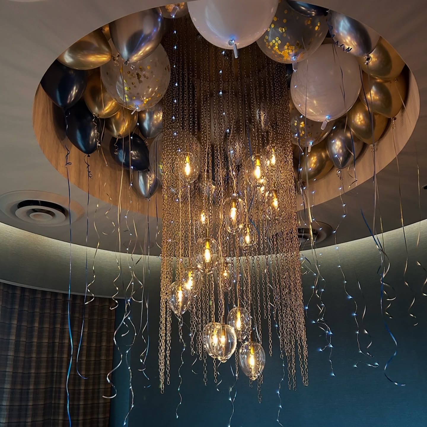 Chandelier surrounded by balloons; silver, gold, and white, hanging from a circular ceiling feature.
