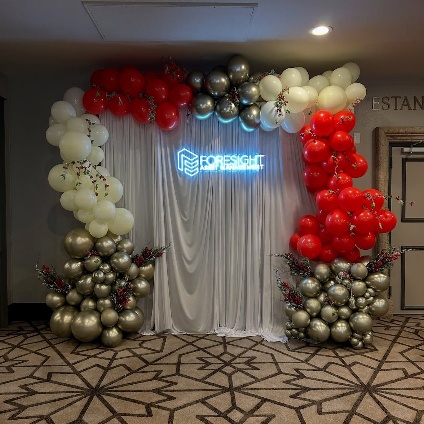 A decorated backdrop with balloons in white, red, and silver, surrounding a neon sign.