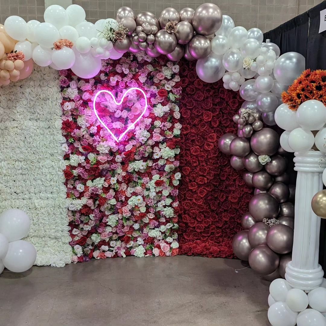 Photo booth with floral walls, neon heart, and balloon arch in pink, white, and red; an event setting.