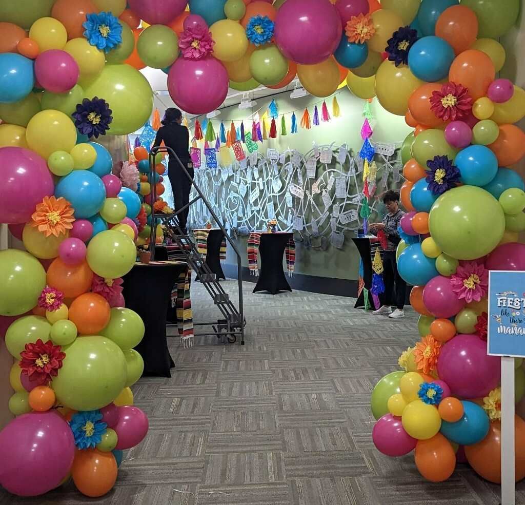Entrance archway decorated with colorful balloons and flowers for a festive event; people in background.