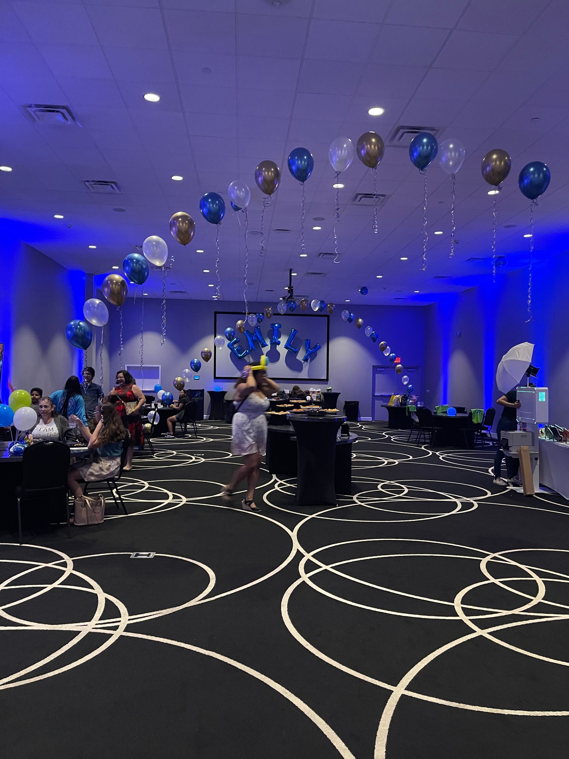 Party in a ballroom with balloons, people, and a projector screen.