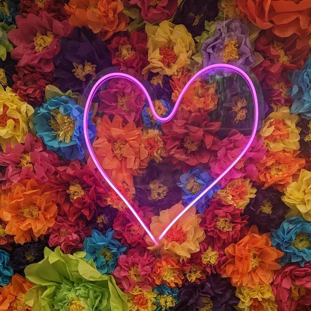 Neon pink heart against a vibrant wall of colorful flowers.