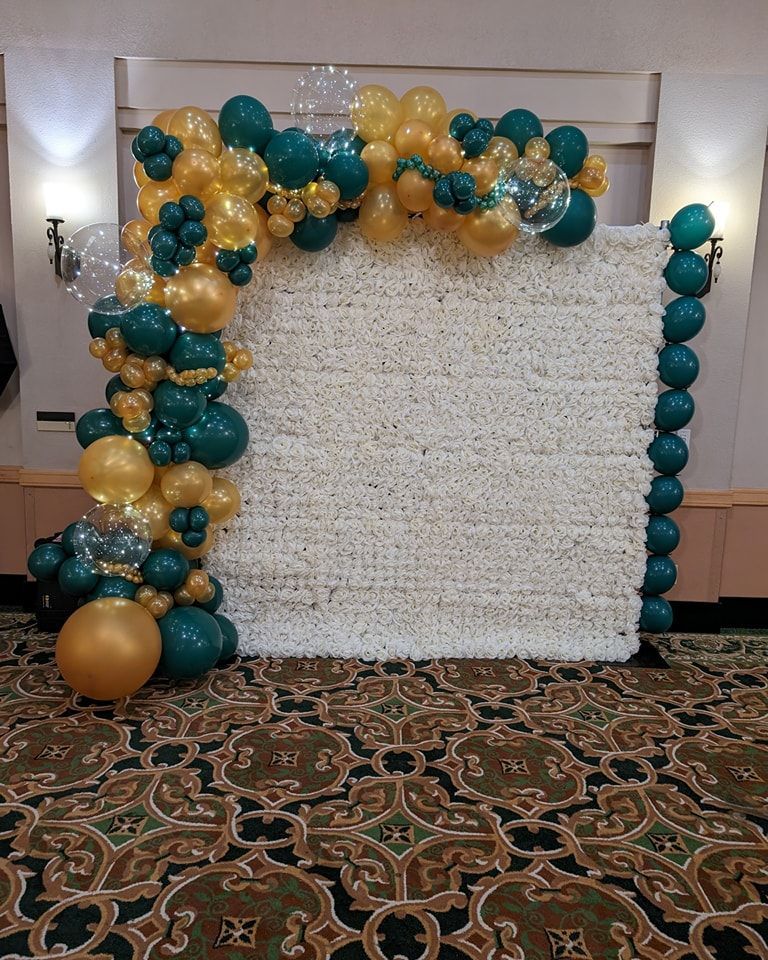 Green and gold balloons adorn a white floral wall, backdrop for an event.