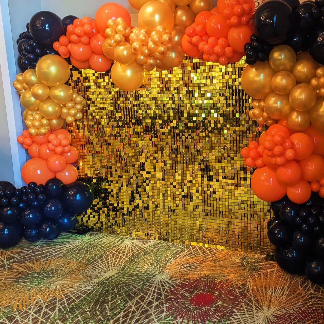 Gold sequin wall with orange, black, and gold balloon arch. Party setting on patterned carpet.