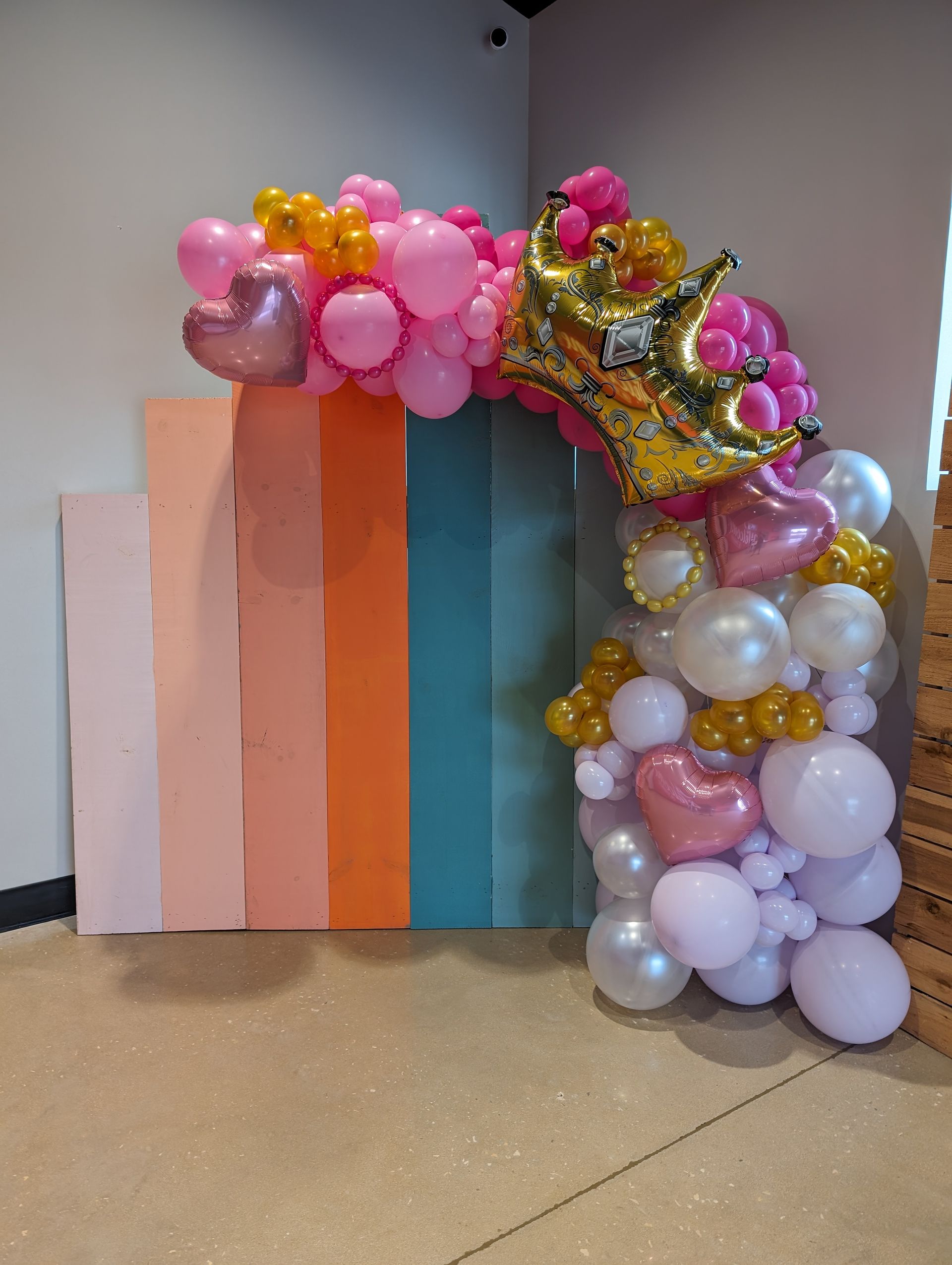 Pink, gold, and white balloon arch with a crown and heart accents, beside a colorful backdrop.