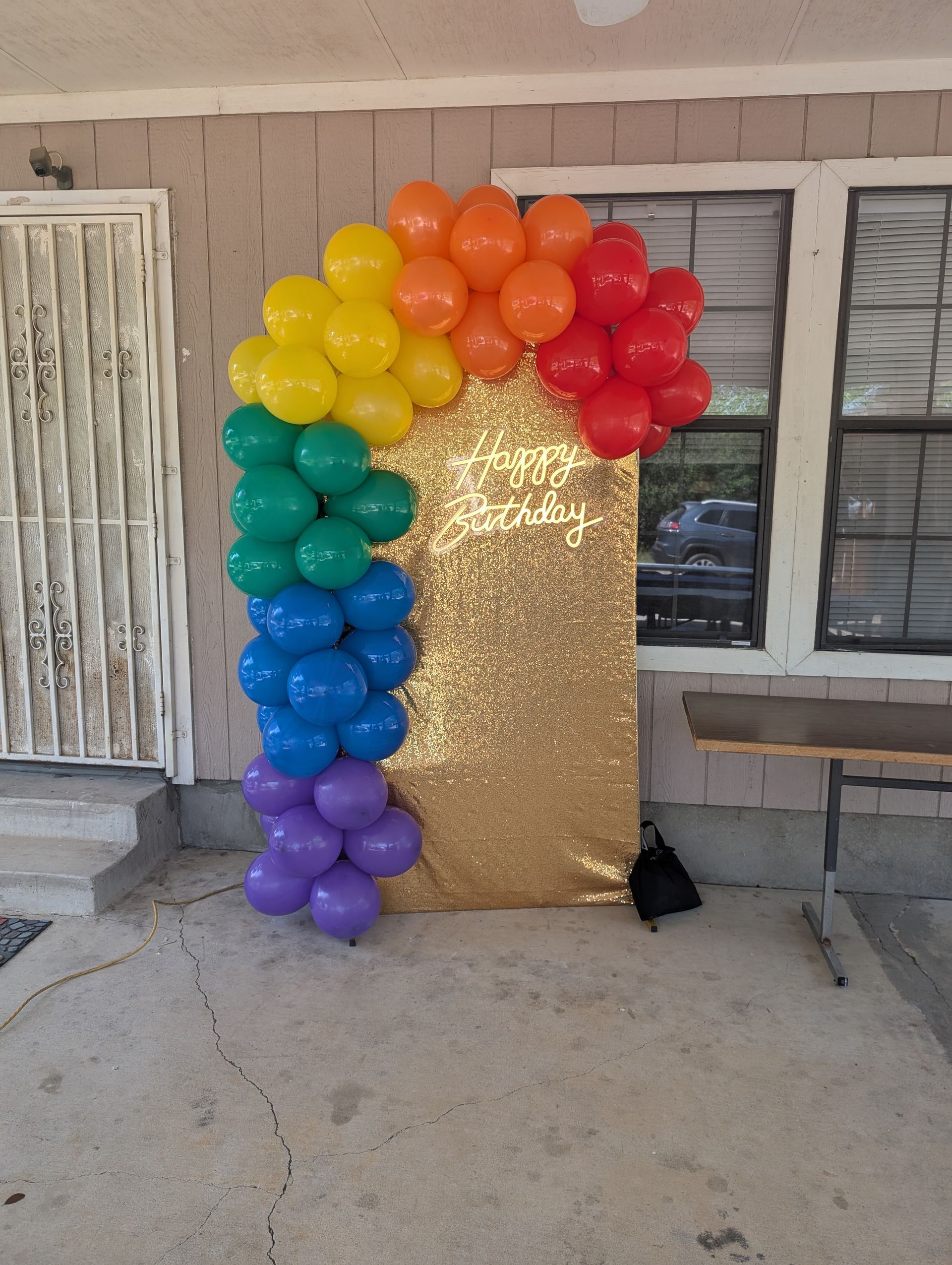 Rainbow balloon arch over a gold sequin backdrop, outside by a building.